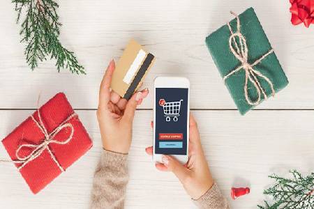 One hand holding a card and one holding a mobile with shopping app. Gifts wraps placed beside it.
